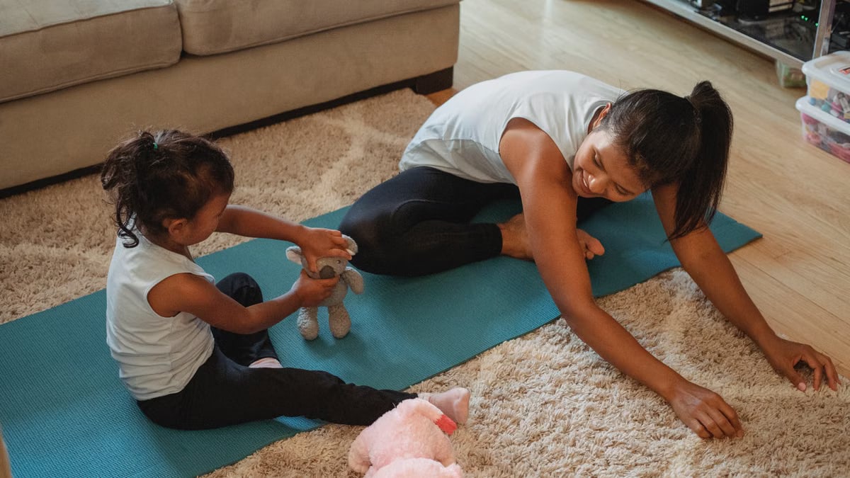 Smiling Hispanic female in activewear stretching on stretch mat and looking at daughter playing with soft toy